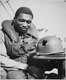 Schwarzweißfoto eines afroamerikanischen Angehörigen der US-Navy während des Zweiten Weltkriegs. Der Soldat in Schwimmweste sitzt an Deck. Er blickt lächelnd auf seinen vor ihm (auf einer Art Beistelltisch) liegenden Stahlhelm, der eine große Delle und ein beinahe faustgroßes Loch mit scharfen, eingerissenen Kanten aufweist. In der originalen Bildunterschrift ist vermerkt, dass die Beschädigung durch einen Schrapnellsplitter entstand, der Soldat aber nur einen oberflächlichen Kratzer erlitt.