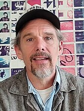 A Caucasian male aged 52 wearing a cap, with grey hair sticking out, and and greying goatie beard, wearing a grey jacket, light blue shirt and white undershirt and piercing hazel eyes