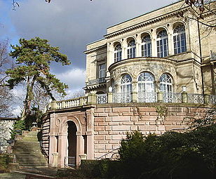 Seitliche Farbfotografie in der Untersicht eines Gebäudes mit einer Terrasse auf einer hohen, runden Mauer mit einer Rundbogenöffnung. Eine verwitterte Treppe führt links an der Mauer vorbei zum Gebäude, das einen runden Vorbau hat. Alle hohen Rundbogenfenster haben Sprossen.