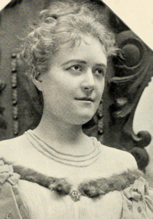 A young white woman with fair hair, wearing a fur-trimmed off-the-shoulder gown, with three strands of beads/pearls at her neck. She appears to be seated in a heavy, carved wooden chair.