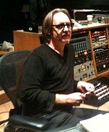 A man wearing glasses and black clothes sits in the control room of a recording studio.