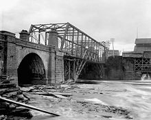 Black and white photo with a stone-arch bridge section in the foreground, and a steel truss bridge spanning river rapids in the background