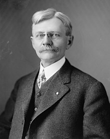 Head and shoulders of a sixtyish man, with a serious expression behind his pince-nez. He has a bushy mustache and his light-colored hair is parted near the top. He wears a three-piece suit, a high collared shirt, and a necktie.