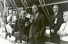 Agnew, standing, speaks into a microphone as others seated in the row of spectators nearby look on.