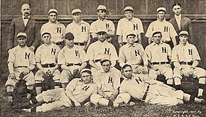 A black and white photograph of seventeen men arranged in three rows. Most are wearing white uniforms with a dark "N" on the chests, but two are wearing dark suits.