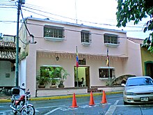 A pink painted adobe building, with Venezuelan flags in front and large plants lining the low wall of a small overhung patio.