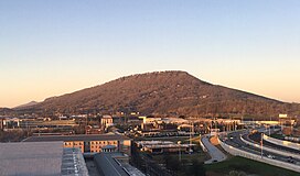 Central tree covered mountain rising up agains a light sky with modern buildings and roads in foreground