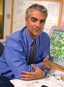 Nicholas Christakis sitting behind desk, wearing light blue shirt, gray patterned tie, and dark blue jeans, looking directly at camera with slight smile