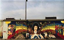 Frontale Farbfotografie von der Berliner Mauer mit einem Bild von einem Mann mit langen, schwarzen Haaren und einem Schnurrbart. Er hält seine Arme schützend vor die Erde, um Kriegswaffen und Industrie fernzuhalten. Aus der Erde wachsen zwei Bäume, die in zwei Regenbögen übergehen, auf denen Menschen tanzen und sich umarmen. Am oberen Bildrand ist das dunkle Universum mit den Planeten zu sehen.