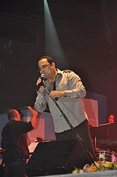 A man wearing glasses and a grey shirt holding a microphone.