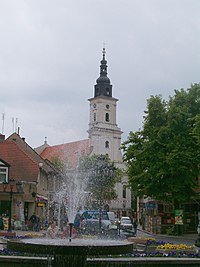 W oddali biały kościół pomiędzy budynkami na końcu ulicy, wieża wybija się ponad dachami. Po prawej stronie drzewa, po lewej stronie kamienice, na pierwszym planie tryskająca fontanna otoczona kwiatami.