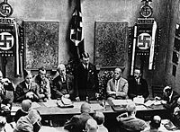 Adolf Hitler (ayakta) 1925'in Şubat ayında NSDAP'nin yeniden kuruluşu vesilesiyle bir konuşma yapıyor. Gregor Strasser ve Heinrich Himmler sağ tarafında ve sol tarafından Franz Xaver Schwarz, Walter Buch ve Alfred Rosenberg bulunmaktadır. Hitler'in arkasında Blutfahne (kan bayrak) yer almaktadır.
