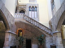 A view of a courtyard inside a building with gray stone arches to right and left and a staircase ascending from right to left across the back wall with potted plants under the stairs. Above the stairs are windows and doors of gothic design.