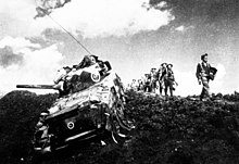 A tank of World War II vintage, with some foot soldiers marching alongside