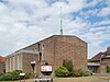 A bulky, plain brown brick building. A flat-roofed projection on the near side has four evenly-spaced ground-to-roof windows with concrete details; the main part of the building behind it has evenly-spaced windows in concrete surrounds, the rightmost of which is much taller than the others. A tall, very thin green metal spire sits on the right side of the roof.