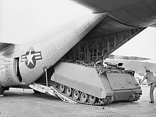 A tracked military vehicle backing onto a ramp at the rear of a large aircraft