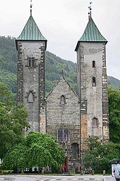 Stone Romanesque church with two towers and a lower, pitched roof entrance hall between them. The towers have slightly curved bronze roofs.