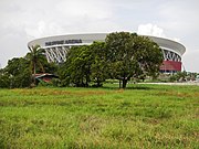 A large stadium with the name "Philippine Arena" displayed on its facade, surrounded by lush green grass and several trees in the foreground. The sky is partly cloudy.