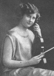 A young white woman, seated, holding a violin upright in her lap, and a bow in her right hand. She has short wavy hair, and is wearing a light-colored satiny dress with no sleeves.