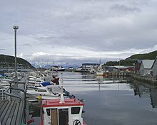 Foto mehrerer kleinerer Boote in einem Hafen, teils umgeben von Häusern. In etwas Entfernung verschneite Berge.