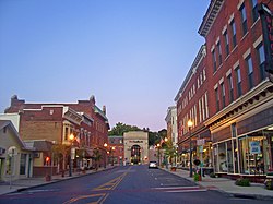 A view of an urban street at dusk with brick buildings on either side and a brown building with a large arched window in the distance at dusk, with the street lamps lit