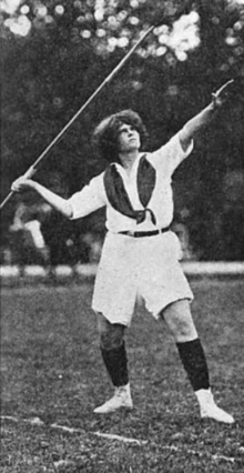 A young white woman with short dark curly hair, about to launch a javelin; she is wearing white athletic clothing, including shorts and a middy blouse