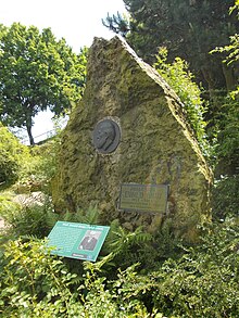 Etwa lebensgroßer oben spitz zulaufender Gedenkstein mit mittig angebrachter Darstellung des Gesichts und mit gesonderter Schrifttafel am Fuße des Steines in begründeter Umgebung.