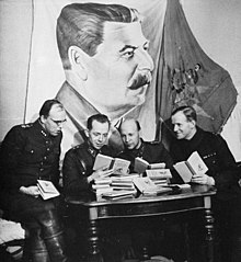 Four Finnish officers in uniform are sitting and reading Soviet skiing manuals with relaxed looks on their faces. A pile of the books is in front of them on a table and a large drape of Joseph Stalin hangs above their heads on the wall.