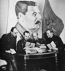 Four Finnish officers in uniform are sitting and reading Soviet skiing manuals with relaxed looks on their faces. A pile of the books is in front of them on a table, and a large drape of Joseph Stalin hangs above their heads on the wall.