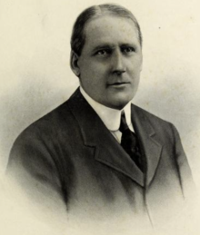 Black-and-white posed photo of Gorman Jr wearing a dark suit coat, darker tie, and a white collared shirt