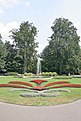 Das Bild zeigt einen Park mit einem Springbrunnen, vor dem sich eine Wiese mit einem Ornament aus Blumen befindet. Im Hintergrund sind hohe Bäume zu sehen.