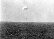 A conical black spacecraft falling towards the surface of the ocean under a single white parachute, seen from some distance away. Very little detail can be seen.