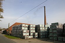 Auf dem Foto ist das Wittmunder Klinkerwerk zu sehen. Im Vordergrund steht eine große Menge verpackter Ziegelsteine auf Paletten. Dahinter ist das Ofenhaus und der Schornstein zu erkennen.