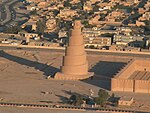 A photograph of a spiral minaret in an open plain with a large number of buildings in the background and a swerving road to the left