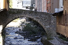 A small stone bridge over a stream of water. Buildings can be seen in the background.