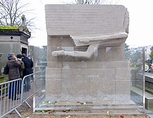 Grabmal Oscar Wildes auf dem Friedhof Père Lachaise in Paris. Das Grabmal besteht aus einem quaderförmigen Sockel, der obere Teil stellt ist eine stilisierte geflügelte Figur im Flug dar. Das Grabmal ist von einer Schutzscheibe umgeben, auf der linken Seite zusätzlich durch Gitter abgesperrt. Jenseits des Gitters stehen vier Menschen. Vor dem Grab liegen mehrere einzelne Blumen. Links und rechts sind die angrenzenden Grabmonumente zu sehen.