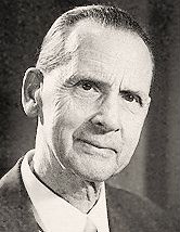 Clean-shaven, elderly man in suit and tie, with short hair, oiled and brushed back, head slightly cocked to one side, half-smiling