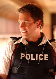 A man with short hair wearing a police bulletproof vest over a button-down shirt.