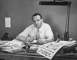 Black and white portrait of a man sitting at his desk, working. The desk is cluttered with books and newspaper and has a large lamp.