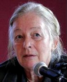 Colour Press shot of an elderly woman with strong features, no makeup, and long, straight white hair, tied back. She is speaking into a microphone.