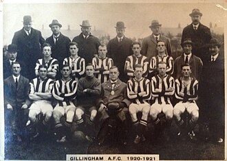 A football team comprising ten players in striped shirts and one in a shirt of a single colour pose for the camera. Five of the men are standing and the other six seated in front of them. Also posing with them are an elderly man in a bowler hat with a chain of office around his neck, and twelve men in business suits, some of whom are wearing hats. A crowd of spectators is visible behind the group.