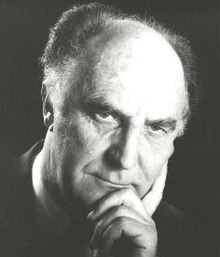 Dramatic publicity shot of a clean-shaven, elderly man with receding hair, with strong features and piercing eyes, looking into the camera, with chin resting on hand.