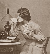 Young white woman in 1914 daywear, seated at a table, teacup in hand