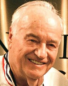 Colour press shot of an elderly, smiling man with short, receding white hair.