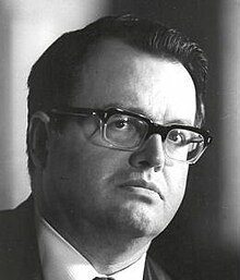 Three-quarter profile publicity shot of a clean-shaven man with serious expression, spectacles, and black hair combed back.