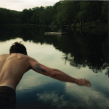 Shows the back of Shawn Mendes as he jumps into a lake surrounded by trees