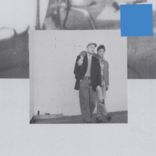 A black-and-white photograph of two men with one's arm around the other set against a larger indistinct photograph, with a blue square in the top-right of the image.