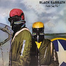 Two pilots stand in front of a fighter jet wearing forest green aviation jumpsuits. They each have odd-looking leather helmets, one coloured tomato red, the other mustard yellow. Their faces are completely covered: Their eyes by dark goggles and their noses and mouths with black leather that has tubes attached for a breathing apparatus.