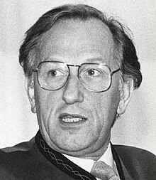 Dramatic, black and white Press photo of a clean-shaven, middle-aged, bespectacled man, with untidy greying hair, brushed back. He is wearing a suit and tie, turning his head, and speaking or singing.