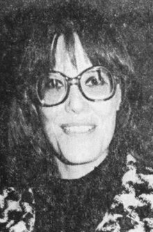 Mary Kenny Badami, a smiling white woman with dark hair cut in a long shag with bangs, wearing eyeglasses and a houndstooth-checked jacket